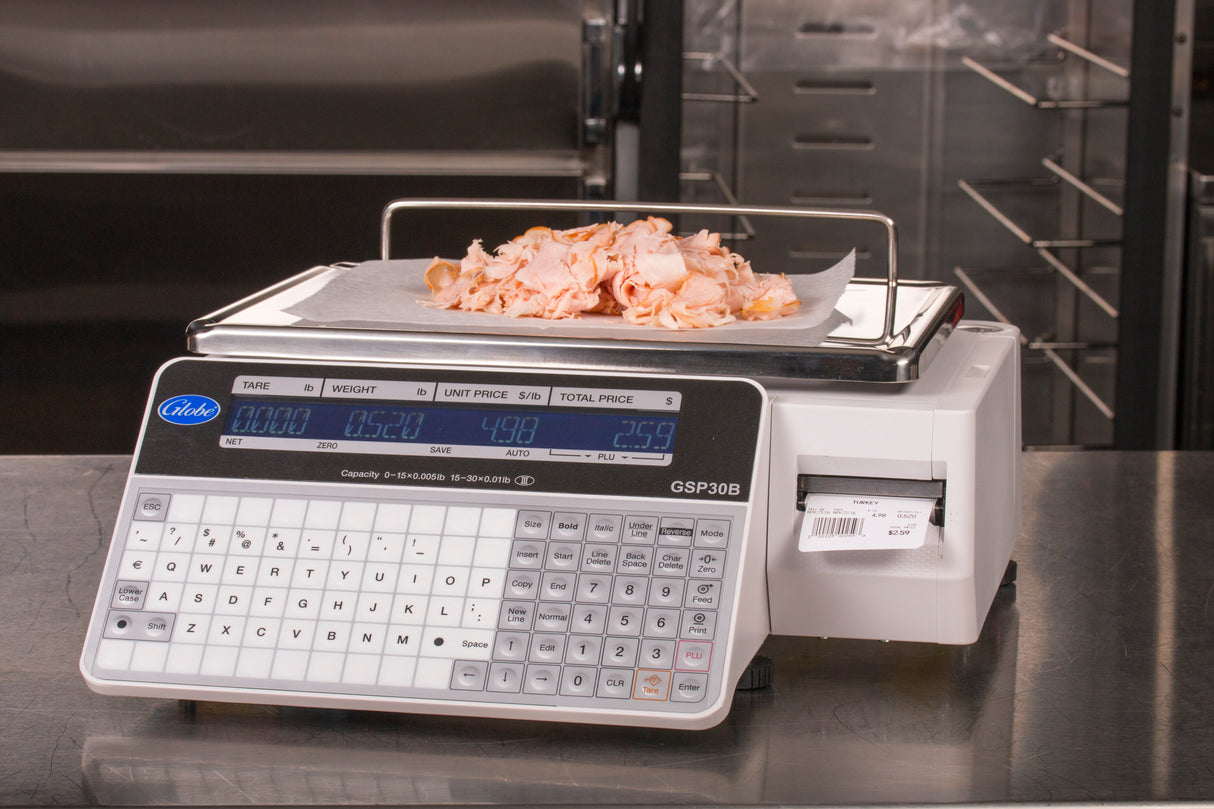 A legal for trade digital deli scale with a keypad and dual-sided LCD display shows sliced deli meat on a parchment-lined tray. The label printing scale displays weight, unit price, total price, and prints a label amid stainless steel equipment.