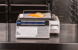 A legal for trade digital deli scale displays 0.565 pounds with sliced cheese on parchment atop its tray. Its keypad, dual-sided LCD display, and label printing scale feature are set against stainless steel surfaces and racks.