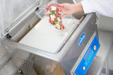 A person in a white lab coat places a vacuum-sealed bag with seasoned raw meat and herbs into a stainless steel Vacuum Packaging Machine labeled “Vak,” which features a blue control panel with buttons, pressure gauge, and gas flush settings on the front.