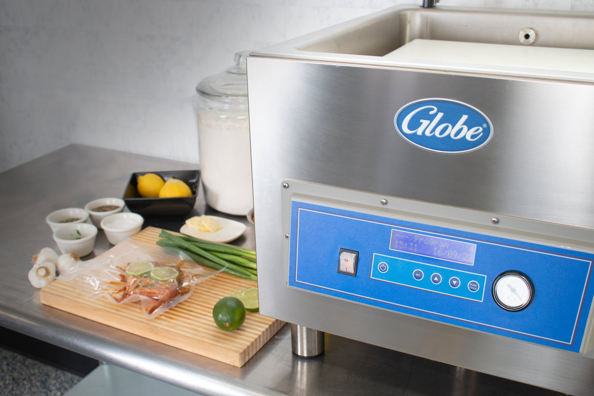 A large stainless steel Globe-brand Vacuum Packaging Machine sits on a counter. Next to it are herbs, lime, sauces, onions, lemon, butter, and flour—ideal for marination programming—all arranged neatly in a clean kitchen setting.