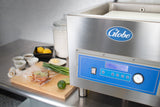 A large stainless steel Globe-brand Vacuum Packaging Machine sits on a counter. Next to it are herbs, lime, sauces, onions, lemon, butter, and flour—ideal for marination programming—all arranged neatly in a clean kitchen setting.