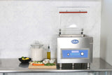 A stainless steel Vacuum Packaging Machine with the lid open sits on a metal counter. Surrounded by a jar of flour, citrus fruit, olive oil, and fresh produce, it features marination programming. The background is a light marble-patterned wall.