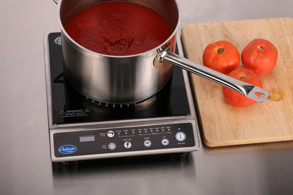 A pot of red tomato sauce heats on a sleek induction range. Nearby, three whole tomatoes sit on a wooden cutting board atop a stainless steel countertop, with the cooktop display and buttons clearly visible in the foreground.