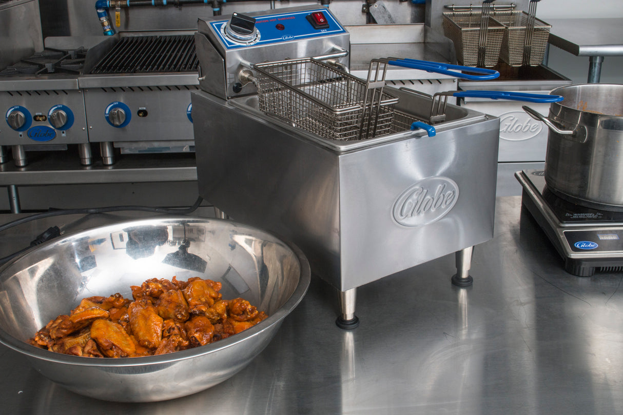 A commercial kitchen with a Globe PF16E electric countertop fryer, 16 lbs. oil capacity, stainless steel basket inside, and a bowl of cooked chicken wings on the metal counter. Stovetop, grill, and fryer baskets are organized on clean surfaces in the background.