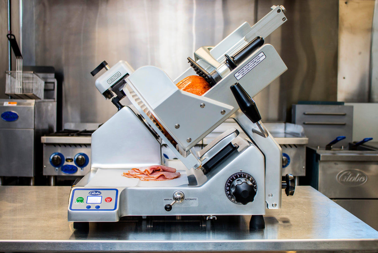 A commercial premium heavy duty slicer with a 13 inch steel knife sits on a stainless steel countertop, slicing deli meat. Sliced pieces rest on the tray below, set against the backdrop of a professional kitchen with stoves and equipment.