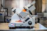A commercial premium heavy duty slicer with a 13 inch steel knife sits on a stainless steel countertop, slicing deli meat. Sliced pieces rest on the tray below, set against the backdrop of a professional kitchen with stoves and equipment.