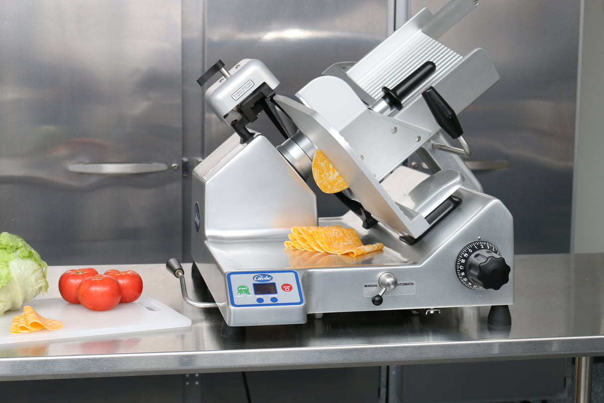 A premium heavy duty slicer sits on a stainless steel counter, with slices of yellow cheese positioned on the slicer tray. Next to it, a cutting board holds tomatoes and lettuce. Metallic cabinets are in the background.