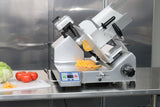 A premium heavy duty slicer sits on a stainless steel counter, with slices of yellow cheese positioned on the slicer tray. Next to it, a cutting board holds tomatoes and lettuce. Metallic cabinets are in the background.