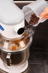 A hand pours chocolate chips from a clear measuring cup into a 5 qt mixer bowl filled with eggs, flour, and other ingredients for cookie dough. The white planetary mixer has a transparent splash guard and sits on a dark countertop.