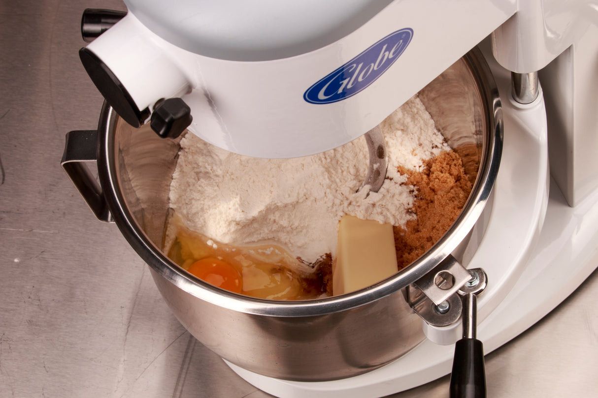 A 5 qt planetary mixer with a metal bowl holds flour, brown sugar, a stick of butter, an egg, and liquid vanilla on a stainless steel counter. The white mixer head displays a blue “Globe” logo, ready to mix dough.