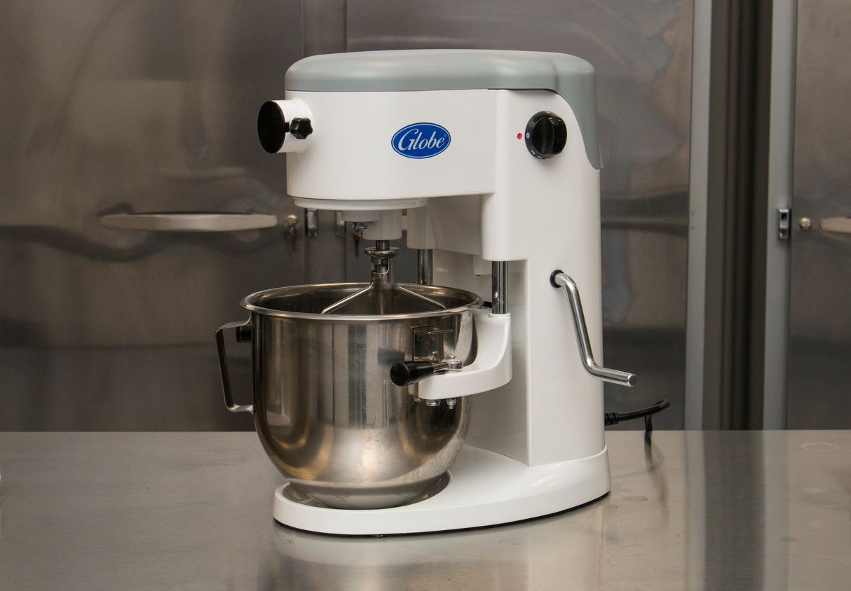 A white Globe brand planetary mixer with a large stainless steel mixing bowl sits on a reflective metal countertop in a commercial kitchen. The countertop mixer features control knobs, a mixing attachment, and metallic cabinets in the background.