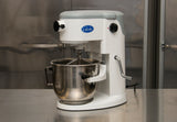 A white Globe brand planetary mixer with a large stainless steel mixing bowl sits on a reflective metal countertop in a commercial kitchen. The countertop mixer features control knobs, a mixing attachment, and metallic cabinets in the background.