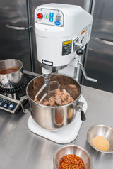 A commercial 8 qt mixer with shredded meat in the bowl sits on a stainless steel counter. Nearby are bowls of red pepper flakes, breadcrumbs, and a pot of sauce on a portable burner, all set in an industrial kitchen setting.