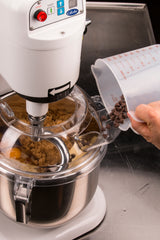 A person pours chocolate chips from a plastic measuring cup into a countertop mixer bowl with brown sugar and eggs. The 8 qt mixer, on a stainless steel counter, is running with its lid partially open to add the ingredients.