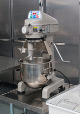 A large 20 qt planetary mixer with a stainless steel bowl and flat beater attachment sits on a metal table in a commercial kitchen, surrounded by stainless steel surfaces and cabinets. A control panel with buttons and a display is on top.