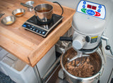 A 20 qt mixer with shredded meat inside sits on a counter next to a wooden prep table holding a hot plate with a pot of brown liquid and small metal bowls containing spices, suggesting food preparation in a commercial kitchen.