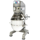A large, industrial commercial floor mixer with a stainless steel mixing bowl, protective wire guard, control panel at the top, and a side crank handle. This gray planetary mixer is designed for heavy-duty food preparation.
