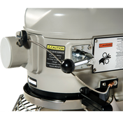 Close-up of a gray industrial machine with a factory installed warning label, metal lever, black knob, and safety cable. The labels highlight caution about gear damage and safety procedures. Part of a protective metal grill is visible at the bottom.