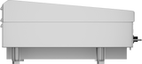 A side view of a modern, minimalist grand piano with a closed lid. The piano is white, has sleek, clean lines, and is elevated on metallic cylindrical legs reminiscent of a polished steel griddle plate. Pedals and controls peek beneath the piano body.