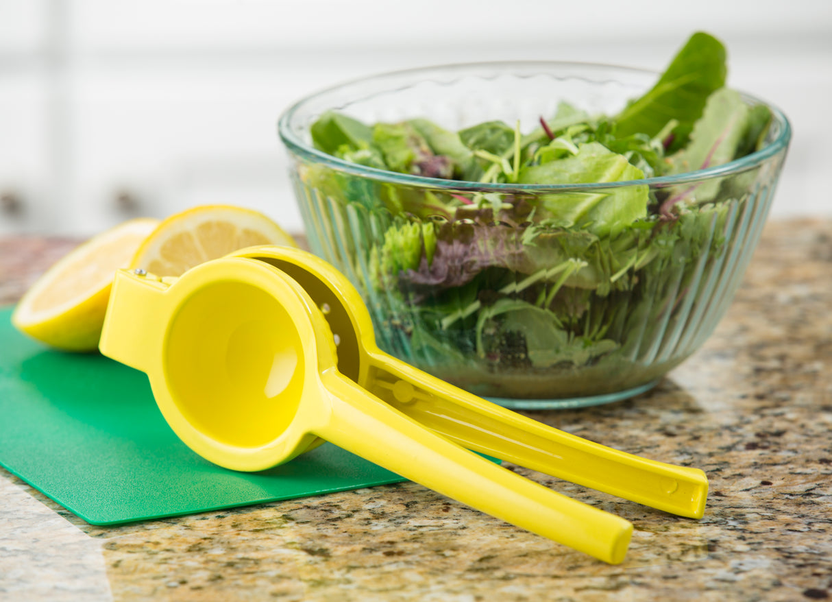 A yellow coated zinc alloy citrus hand squeezer rests on a green cutting board with a halved lemon. Behind them, a glass bowl of mixed leafy greens sits atop a speckled kitchen countertop.