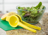 A yellow coated zinc alloy citrus hand squeezer rests on a green cutting board with a halved lemon. Behind them, a glass bowl of mixed leafy greens sits atop a speckled kitchen countertop.