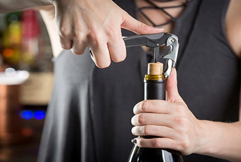 A person uses a waiter's corkscrew to open a wine bottle. One hand holds the bottle steady while the other turns the corkscrew into the cork. The person wears a dark sleeveless top with crisscross laces at the neckline. The background is softly blurred.
