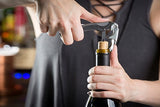 A person uses a waiter's corkscrew to open a wine bottle. One hand holds the bottle steady while the other turns the corkscrew into the cork. The person wears a dark sleeveless top with crisscross laces at the neckline. The background is softly blurred.
