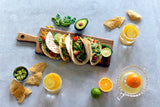 Three tacos with lettuce, cheese, and tomato are on a wooden board, garnished with lime slices. Nearby are tortilla chips, guacamole, sliced jalapeño, a halved avocado, and orange drinks beside juice freshly squeezed with a TableCraft glass juicer.