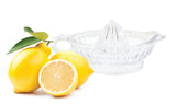 Two whole lemons, one with leaves, and a halved lemon sit next to a clear TableCraft glass juicer. The white background highlights the bright yellow lemons and the transparent juicer beautifully.
