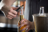 A person uses a stainless steel pocket bottle opener to open a glass bottle of beer, while another hand holds the bottle steady. A wicker-patterned metal ice bucket with a second unopened bottle sits beside them in the softly lit indoor scene.