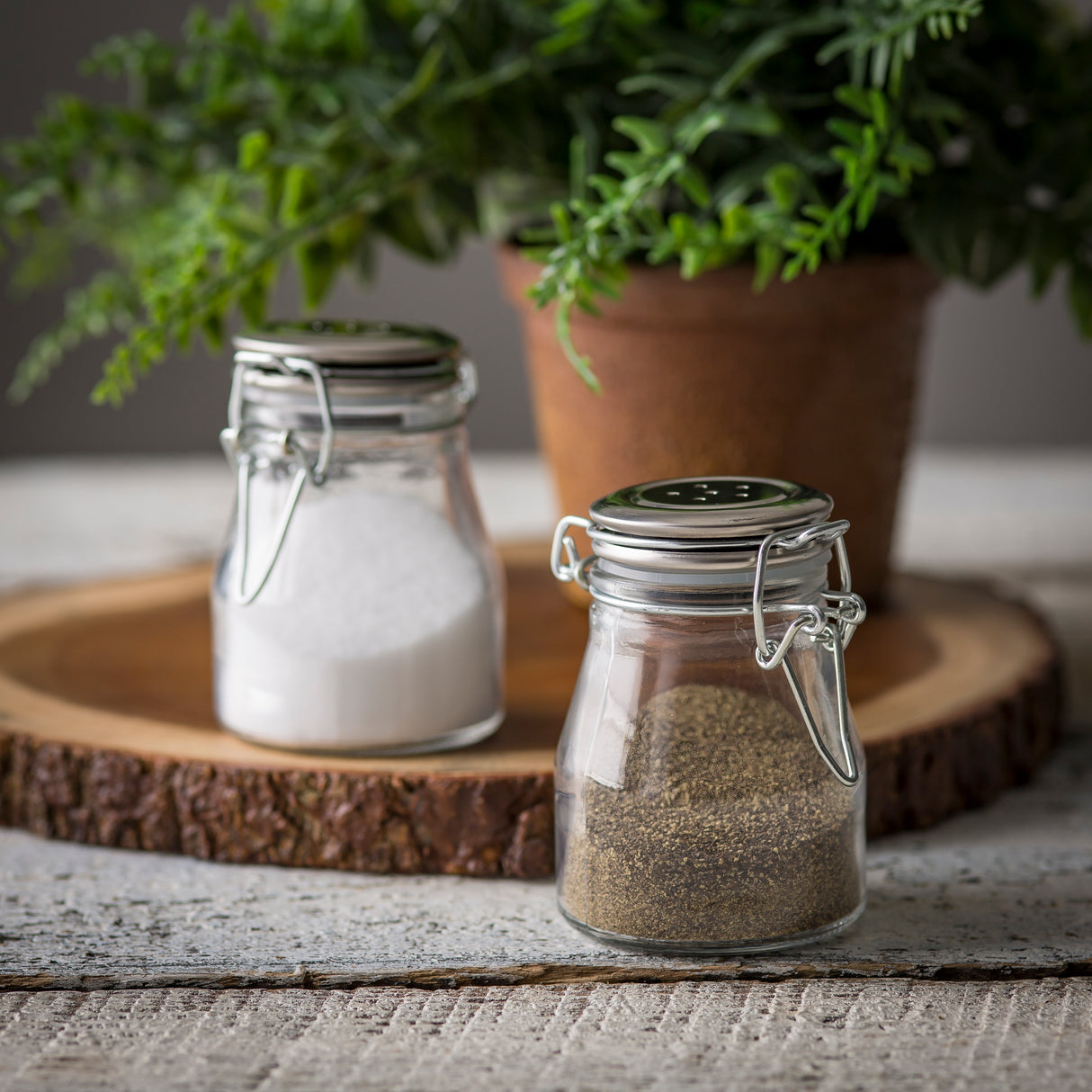 Two small glass shakers with stainless steel clip top clasps, one filled with white salt and the other with ground black pepper, sit on a rustic wooden surface. A wooden tray and a potted green plant add to the cozy kitchen scene.