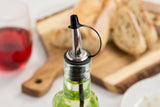 A close-up of a glass bottle with a stainless steel pourer and black tethered cap, filled with green oil. In the blurred background, sliced bread rests on a wooden board next to a glass of red wine, all set on a white surface.