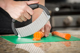 A person uses a crinkle food cutter with a stainless steel blade to slice a carrot on a green cutting board in the kitchen. The hand grips the black handle firmly, and wavy carrot slices are visible near the stove.