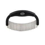 A dishwasher safe crinkle food cutter with a black, curved handle and a wide, wavy stainless steel blade. The blade has vertical grooves for crinkle-cut shapes, and an oval metal logo plate is set into the handle’s center. The background is white.