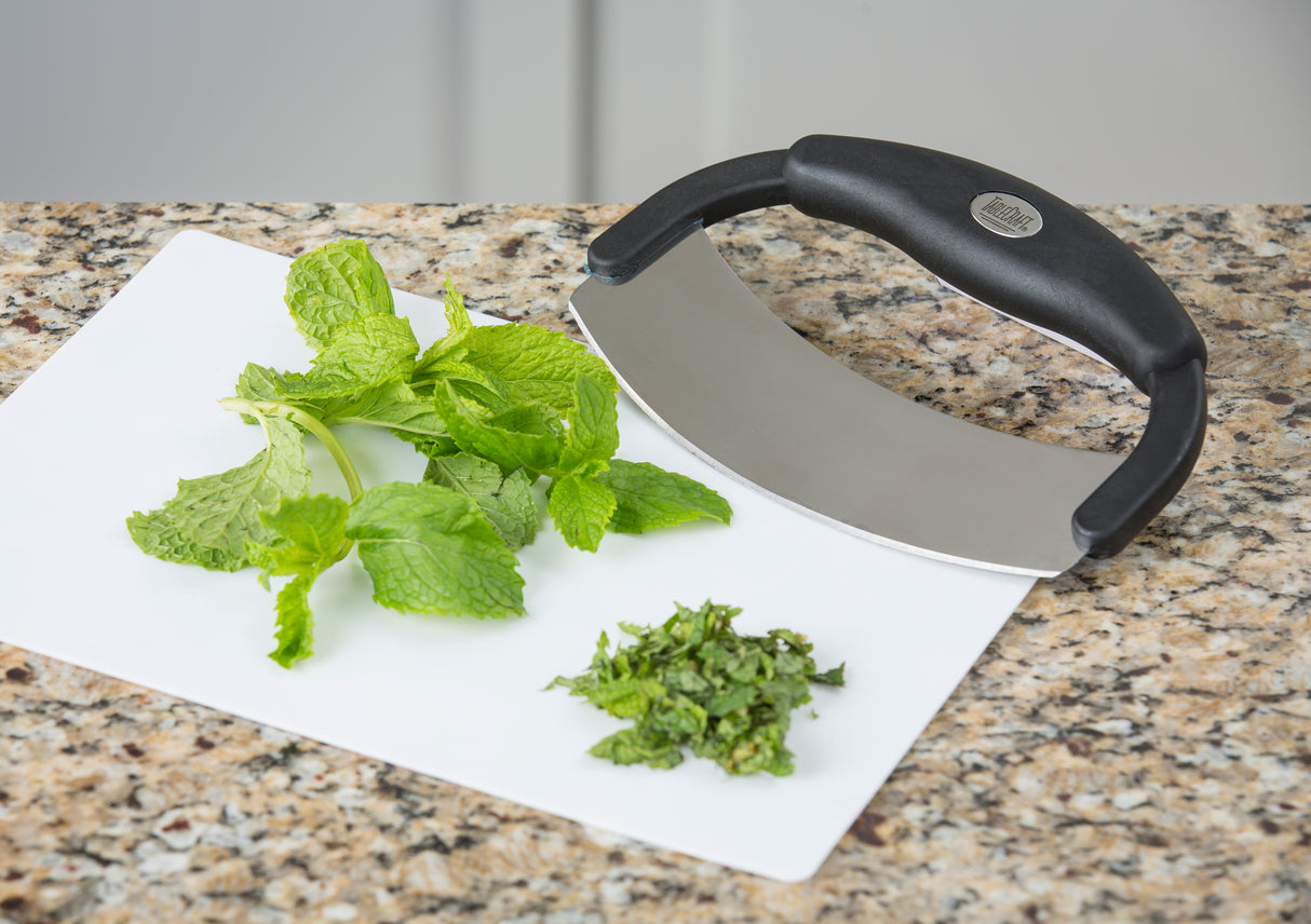 A handheld mezzaluna food cutter with a stainless steel blade and black handle rests on a white cutting board atop a granite countertop. Fresh mint leaves are on the board, some already finely chopped beside the curved blade.