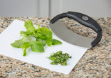 A handheld mezzaluna food cutter with a stainless steel blade and black handle rests on a white cutting board atop a granite countertop. Fresh mint leaves are on the board, some already finely chopped beside the curved blade.