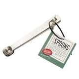 A set of four small stainless steel measuring spoons labeled “SMIDGEN” and attached by a ring, crafted with the quality of J.A. Henckels Steel and a green and white tag reading “Spice Spoons, Set of 4, Stainless Steel, Tablecraft.”.