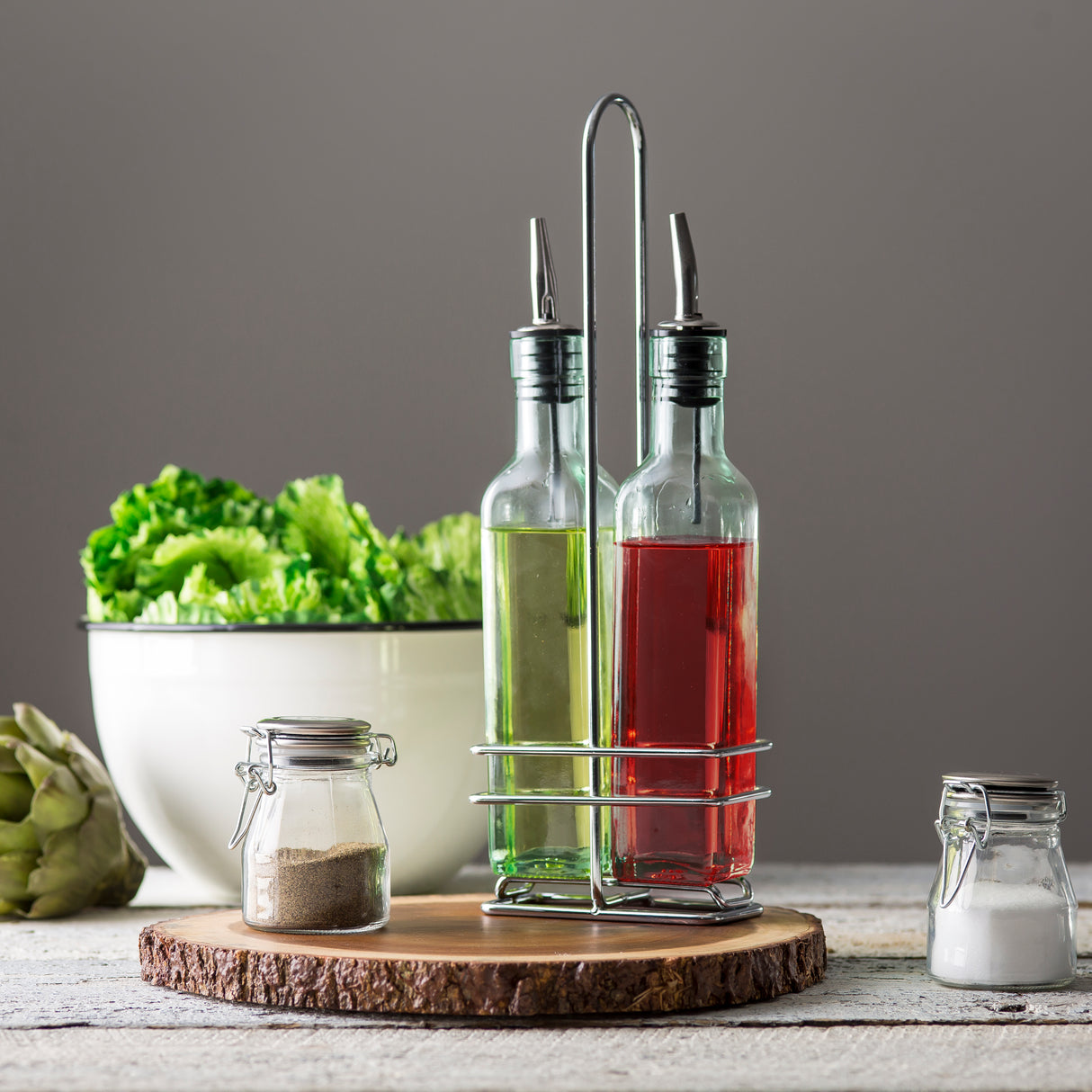 Two green tint glass bottles with stainless steel pourers—one with green liquid, one red—stand in a chrome plated rack on a wooden tray. A bowl of lettuce, an artichoke, and glass salt and pepper jars sit nearby on a rustic white table.