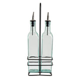 Two TableCraft Products green tint glass bottles with stainless steel pourers are placed side by side in a black metal stand with a handle. The rectangular bottles feature rounded shoulders, and the open stand has a minimalist design against a white background.