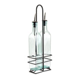 Two clear glass bottles with stainless steel pourers are held upright in a black metal wire holder with a handle. The holder’s rectangular base and grid-like sides keep the bottles secure and organized. The bottles are empty and have a slight blue tint.