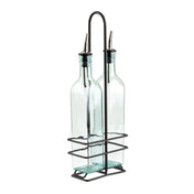 Two clear glass bottles with stainless steel pourers are held upright in a black metal wire holder with a handle. The holder’s rectangular base and grid-like sides keep the bottles secure and organized. The bottles are empty and have a slight blue tint.