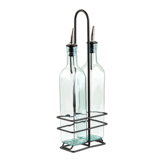 Two clear glass bottles with stainless steel pourers are held upright in a black metal wire holder with a handle. The holder’s rectangular base and grid-like sides keep the bottles secure and organized. The bottles are empty and have a slight blue tint.