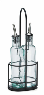 Two clear Gemelli bottle set glass oil and vinegar bottles with stainless steel pourers sit side by side in a black metal holder with a round base, vertical bars, and handle. The background is white.