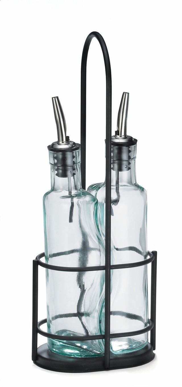 Two clear Gemelli bottle set glass oil and vinegar bottles with stainless steel pourers sit side by side in a black metal holder with a round base, vertical bars, and handle. The background is white.