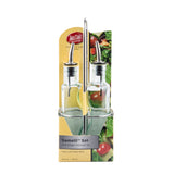 Two glass oil and vinegar dispensers with a green tint, featuring stainless steel pourers, sit in a metal rack. The yellow and green packaging displays a salad image, the TableCraft brand, and reads: Gemelli bottle set, 8.5 fl oz / 250 ml.