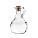 A clear glass bottle by TableCraft Products with a round body, narrow neck, and curved handle, sealed with a brown cork stopper. This elegant Olive Oil Bottle is empty and set against a white background.