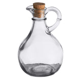 A clear glass olive oil bottle by TableCraft Products with a round body, narrow neck, curved handle, and cork stopper. Empty and elegant, it sits on a white background to showcase its transparency and simple design.