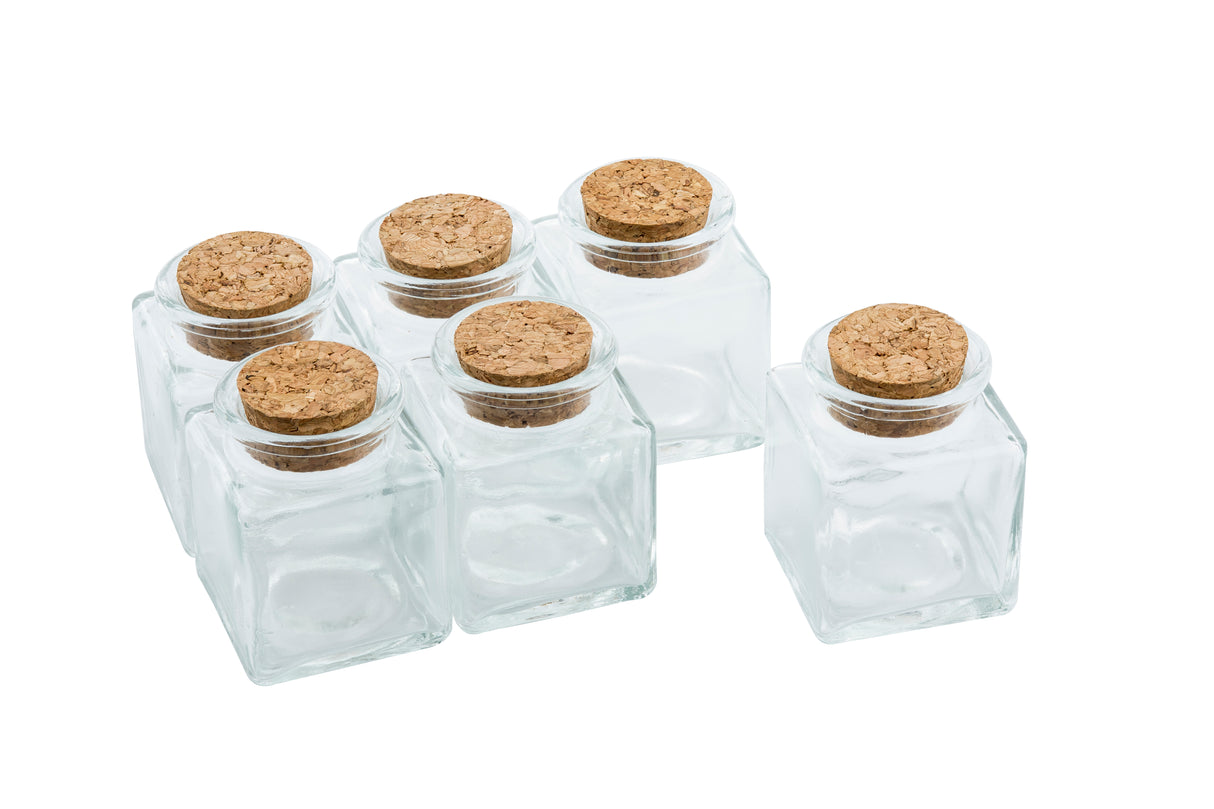 Six small, empty TableCraft mini spice jars with square bases and round openings are arranged closely together. Each glass spice jar is sealed with a round cork stopper and set against a plain white background.