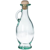 A clear olive oil bottle with a curved body, a small handle on one side, a narrow neck, and a round cork stopper at the top. The empty bottle stands upright on a white background.