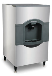 A large, stainless steel Scotsman iceValet hotel ice dispenser with black trim stands on four adjustable legs. The front features a push lever for dispensing ice and a recessed area for cups, with the Scotsman logo above the lever and 180 lb storage capacity.